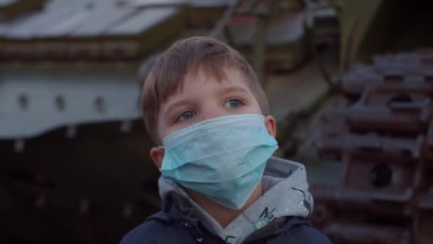 Boy in medical protective mask stands by military machine during pandemic outbreak of coronavirus COVID-19 and watches around. Quarantine emergency and martial law to combat coronavirus. Close up - Materiaali, video