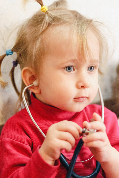 Children's role- playing games. Portrait of a two-year-old girl in a bright pink jacket playing doctor with a stethoscope at home. - Foto, imagen
