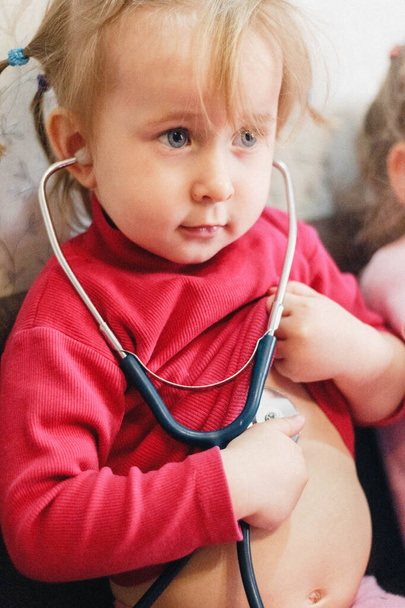 Children's role- playing games. Portrait of a two-year-old girl in bright pink clothes, listening to herself with a stethoscope and portraying a doctor. - Photo, Image
