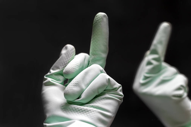 Uma mão em uma luva de borracha verde de hortelã mostra o dedo médio para sua reflexão no espelho na sala preta escura. Vai-te lixar. Foco seletivo. Vista de perto
 - Foto, Imagem