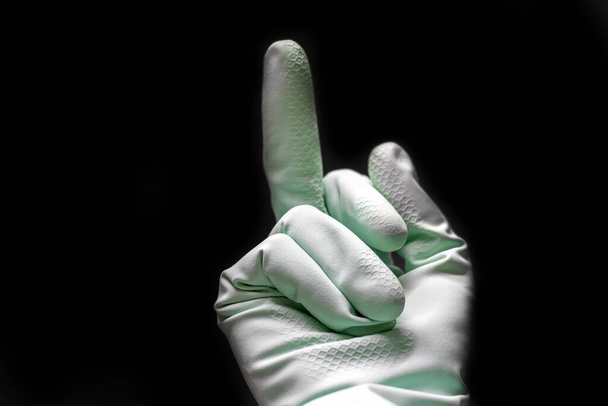 A hand in a mint green rubber glove shows middle finger in the dark black room. Fuck you concept isolated on black background. Selective focus. Closeup view - Foto, afbeelding