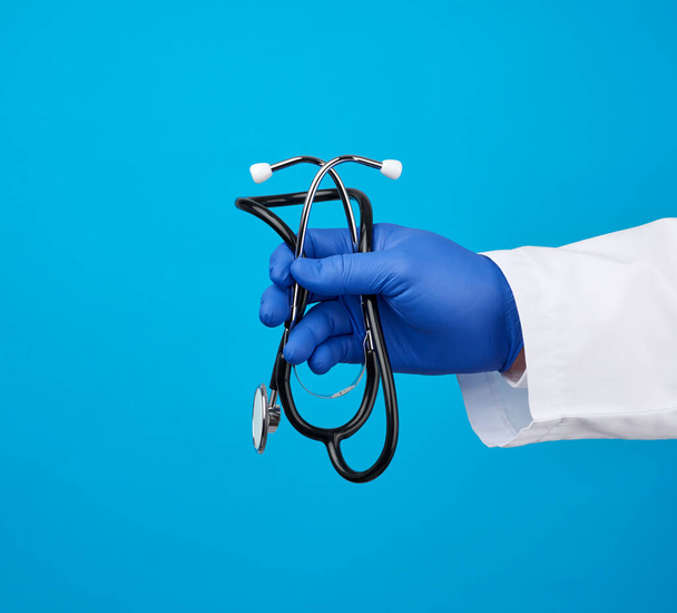 Arzt im weißen Arztkittel hält ein schwarzes Stethoskop auf blauem Hintergrund, Kopierraum - Foto, Bild