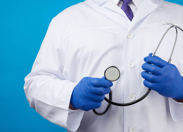 erwachsener Arzt im weißen Arztkittel steht und hält ein schwarzes Stethoskop auf blauem Hintergrund, Kopierraum - Foto, Bild