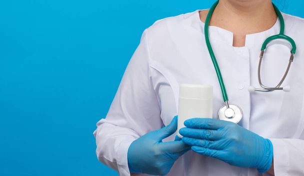 Frauen Hand in blaue sterile Handschuhe hält ein weißes Plastikgefäß für Pillen, medizinisches Behandlungskonzept für Krankheiten, blauer Hintergrund, Kopierraum - Foto, Bild