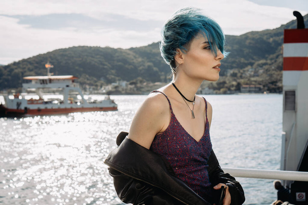 Portrait of a girl with blue hair in a purple dress and a black jacket on a background of a bay by the mountains - Fotografie, Obrázek