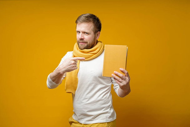 Lindo hombre en una bufanda, sostiene un libro amarillo en las manos, lo señala con el dedo índice y mira a la cámara
. - Foto, Imagen