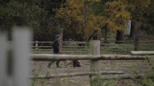 Bobruisk, Belarus - 14 October 2019: A man walks with a wheelbarrow to a cattle ranch along a wooden fence. Side view - Felvétel, videó