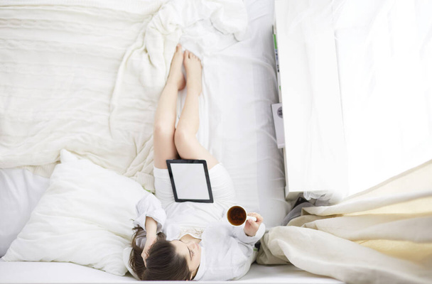 Portrait of woman using tablet and drinking coffee while sitting on the bed in the morning - 写真・画像