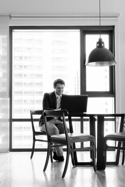 Portrait of young handsome businessman sitting in the dining room near the kitchen in black and white - Photo, Image