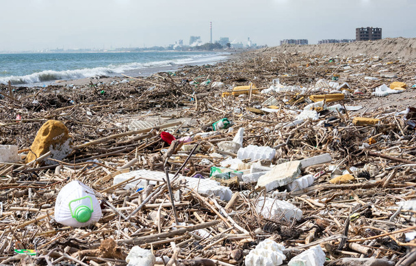 Una spiaggia selvaggia piena di rifiuti e spazzatura, soprattutto plastica, proveniente dall'uomo. Sullo sfondo una fabbrica inquinante e altri edifici
 - Foto, immagini