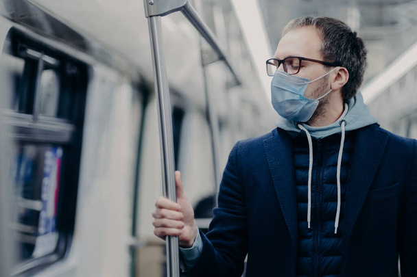 Kranker Mann atmet durch medizinische Maske, fährt mit öffentlichen Verkehrsmitteln, schützt sich vor Coronavirus oder Grippe, bekommt Arbeit in der U-Bahn, trägt Brille und Kapuzenpullover. Pandemiekonzept - Foto, Bild