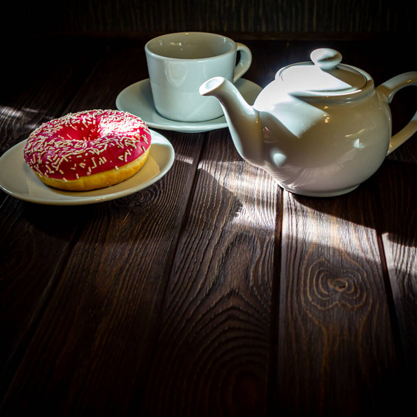 White tea cup, white teapot and donut on a white saucer on a wooden table in the sunlight - Фото, изображение