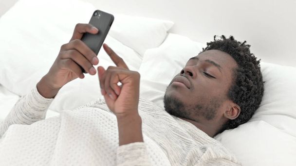 Africano Man Scrolling é Smartphone na cama
 - Foto, Imagem