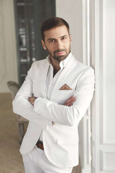 Elegant young handsome man with a beard in a white classic suit. The hotels interior. Photo Studio. Sofa. Window. Chair. Chandelier - Foto, immagini