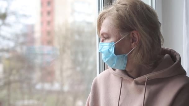 Szomorú nő orvosi maszkkal az ablak mellett. - Felvétel, videó