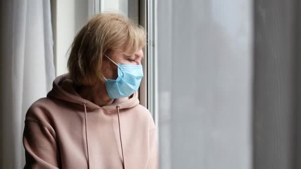 Smutna starsza kobieta w masce medycznej przy oknie - Materiał filmowy, wideo