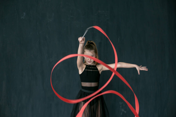 Маленькая гимнастка в черном трико и черная пачка стоят лицом к камере и выполняют упражнение с ярко-красной лентой на темно-сером фоне
 - Фото, изображение