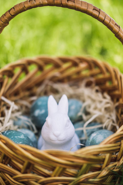 Painted blue textured easter eggs and a white plastic rabbit in a wicker brown hand made basket on green fresh grass. The concept of the spring holiday and egg hunting. - Фото, изображение