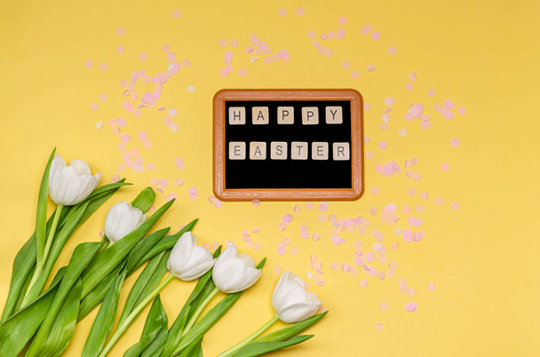 Chalk board with the words Happy Easter, pink decorative paper confetti and a bouquet of white tulips on a yellow background, copy space, flat lay. The concept of the holiday, spring. View from above. - Photo, Image