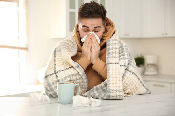 Хворий молодий чоловік з чашкою гарячого напою і тканинами на кухні. Вірус грипу
 - Фото, зображення