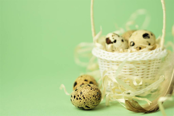 Белая плетеная корзина, наполненная соломой, перепелиными яйцами и перьями на зеленом фоне. Концепция пасхальных праздников. Пасхальная карточка.
. - Фото, изображение