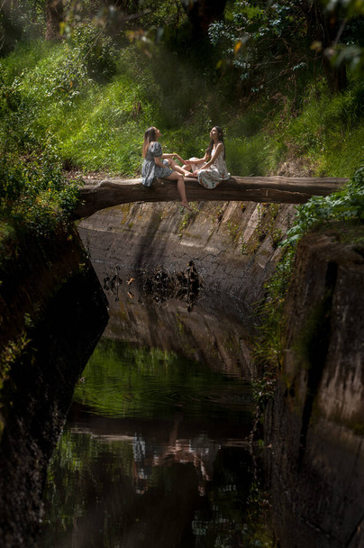 mère et fille assises face à face sur un pont rustique en bois baigné au soleil sur une rivière au milieu de la nature
 - Photo, image