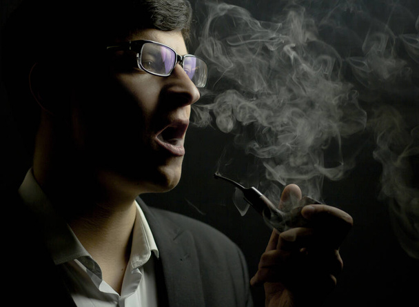 A young brunette smokes a pipe in the dark. Man smoking a pipe - Photo, Image