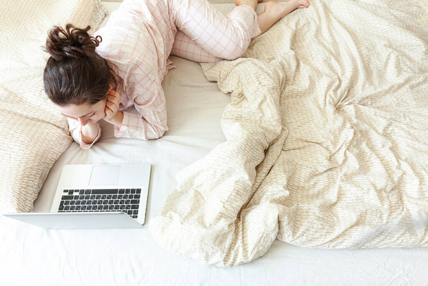 Κινητό γραφείο στο σπίτι. Νεαρή γυναίκα με πιτζάμες κάθεται στο κρεβάτι στο σπίτι και εργάζεται χρησιμοποιώντας τον υπολογιστή του φορητού υπολογιστή. Κορίτσι του τρόπου ζωής σπουδάζει σε εσωτερικούς χώρους. Έννοια ελεύθερης επιχειρηματικής καραντίνας - Φωτογραφία, εικόνα