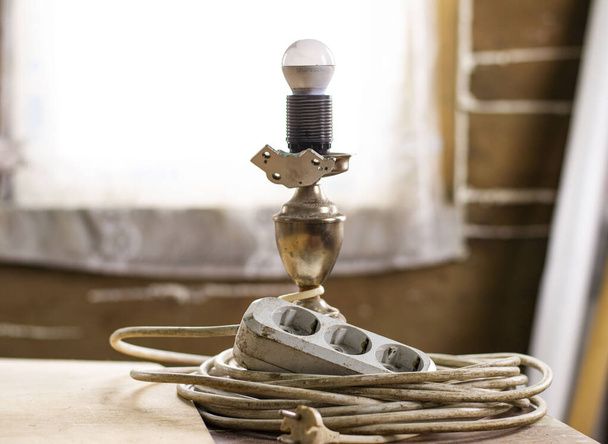 lámpara de mesa con lámpara led y un cable de extensión eléctrica blanca borrosa en la suciedad de construcción. sitio de construcción. reparación en una casa de madera
 - Foto, imagen
