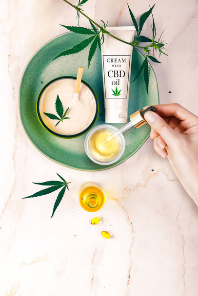 Pipette avec de l'huile cosmétique CBD dans les mains d'une femme sur un fond de table avec des cosmétiques, crème avec du cannabis et des feuilles de chanvre, marijuana
 - Photo, image