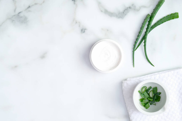 Cosmetic cream jar and green sliced stems aloe vera on marble background. Organic moisturizer hand cream. Hand skin care and beauty treatment concept - 写真・画像