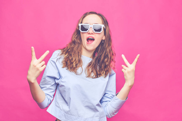 Stylish caucasian girl in a pale blue t-shirt and 8-bit glasses on a pink background. Gesture with two hands - peace. Close up. Toned. - Photo, Image