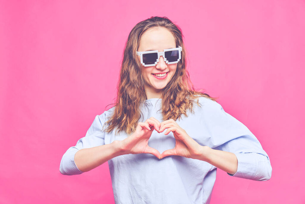 Stylish caucasian girl in a pale blue t-shirt and 8-bit glasses on a pink background. Hand gesture heart, love. Close up. Toned. - Photo, Image