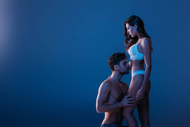 сексуальний чоловік стоїть на колінах і обіймає привабливу дівчину в білій білизні ізольовано на темно-синьому
 - Фото, зображення