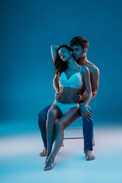 shirtless man and seductive girl in white lingerie sitting on chair and hugging on blue background - Foto, Bild