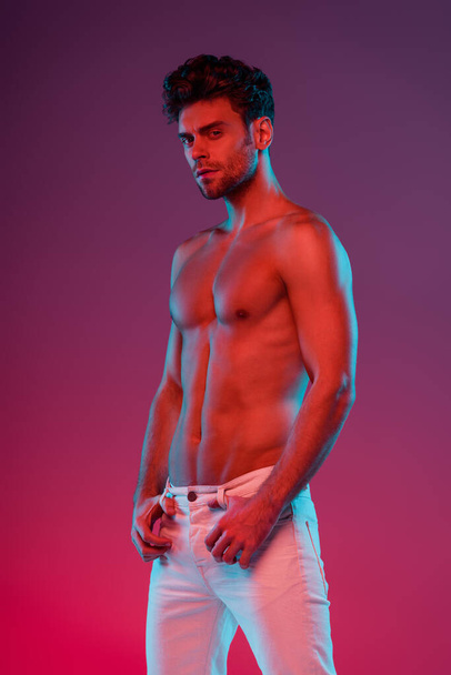 sexy, confident man looking at camera while posing on purple background with gradient - Φωτογραφία, εικόνα