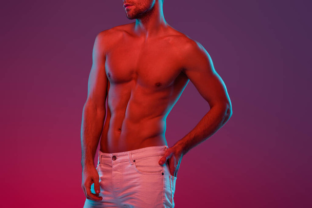 partial view of sexy, shirtless man holding hand in back pocket while posing on purple background with gradient - Zdjęcie, obraz