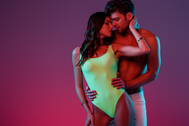 handsome shirtless man hugging sexy girl in swimsuit touching his neck on purple background with gradient - Photo, Image