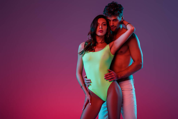 sexy shirtless man hugging attractive girl in swimsuit touching his neck on purple background with gradient - Foto, Imagem