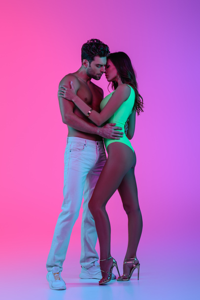 vista completa de hombre sexy sin camisa en jeans blancos y chica seductora en traje de baño verde abrazándose sobre fondo rosa y púrpura
 - Foto, imagen