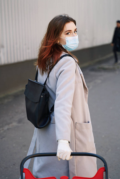 Young woman with beautiful blue eyes wearing protection face mask against coronavirus COVID-2019, Novel coronavirus 2019-nCoV pulling a shopping cart on supermarket department store wall background - Фото, изображение