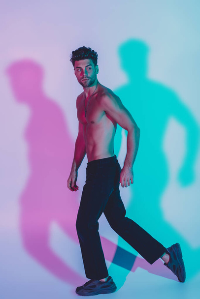 full length view of sexy, shirtless man in dark blue jeans looking back while walking on blue background with shadows - Zdjęcie, obraz