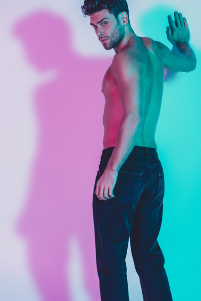 sexy shirtless man in dark blue jeans touching wall while standing on background with blue and violet shadows - Photo, Image