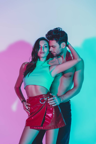 sexy man touching hips of sensual girl embracing his neck and looking at camera on blue background with turquoise and violet shadows - Фото, изображение