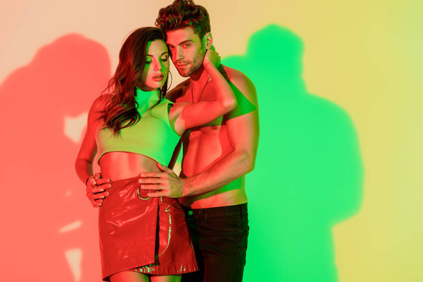 sexy, shirtless man looking at camera while touching hips of seductive girl embracing his neck on yellow background with red and green shadows - Photo, Image