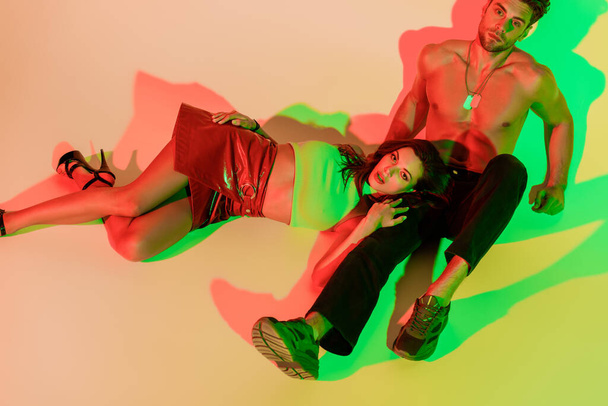 beautiful, stylish girl looking at camera while lying on sexy, shirtless man sitting on yellow background with red and green shadows - Фото, изображение