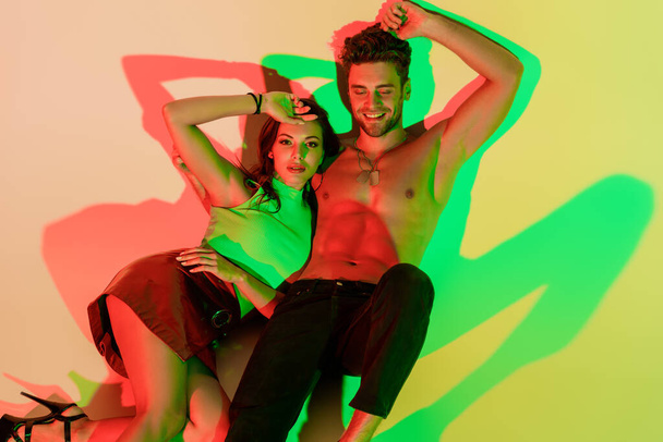 sensual, trendy girl and sexy man smiling while lying on yellow background with red and green shadows - Photo, Image