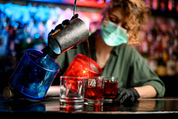 Young bartender girl in medical mask and black gloves energetically pour drink from steel glass into glasses with ice. - Photo, Image