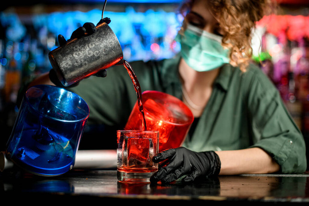 Молодая барменша в медицинской маске и черных перчатках профессионально наливает напиток из стального стекла в стаканы со льдом
. - Фото, изображение