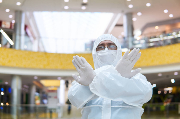 человек в одноразовом противоэпидемическом антибактериальном костюме-изоляторе демонстрирует решительный жест остановки двумя руками на фоне какого-то интерьера общественного пространства
 - Фото, изображение